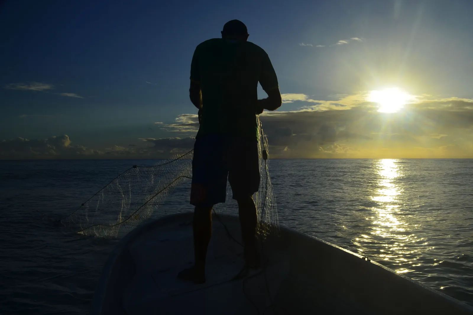 Silhueta de um pescador visto de costas, em pé na canoa, recolhendo a tarrafa contra o sol nascente refletido na água.