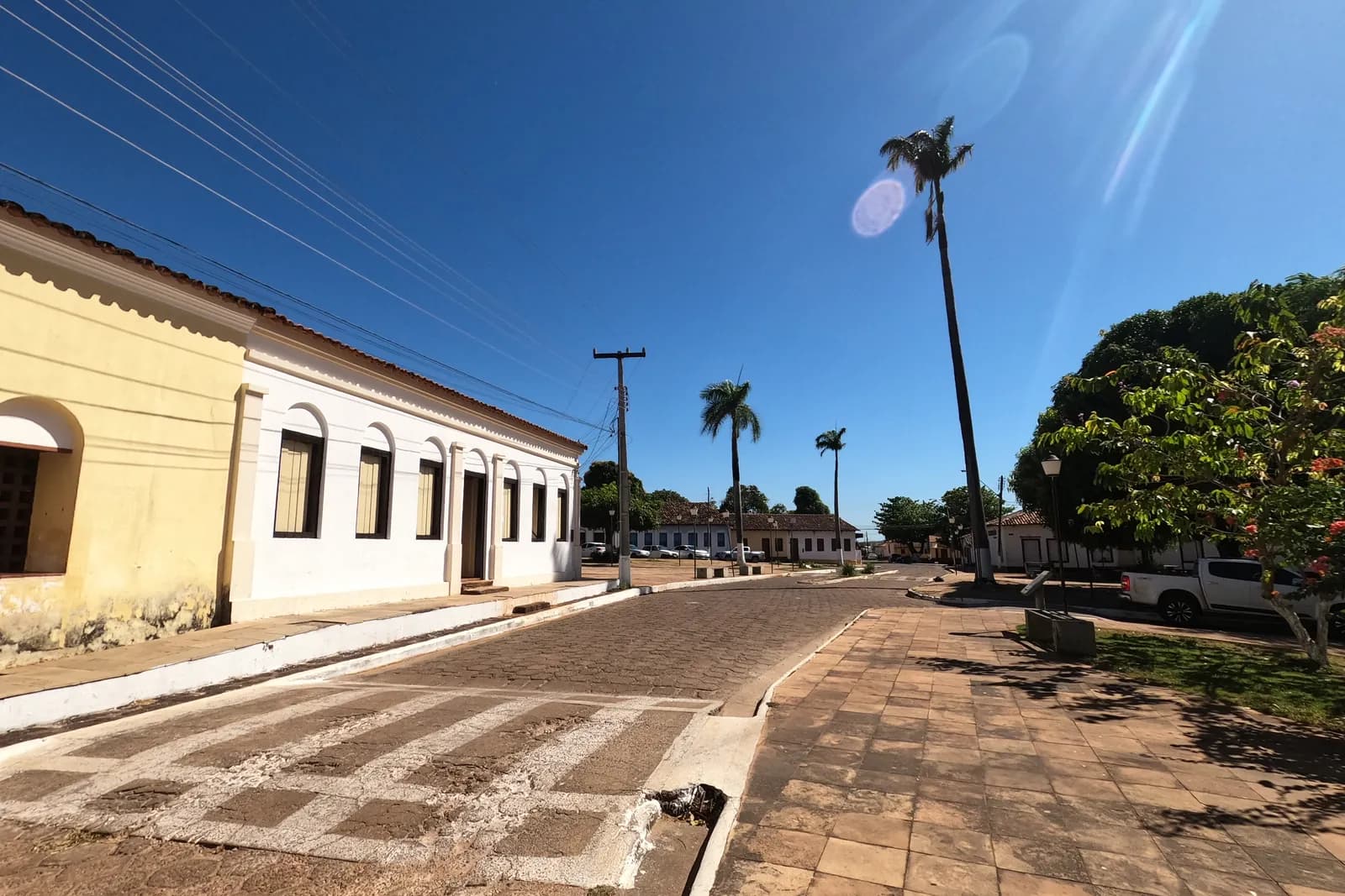 Rua calçada de pedra em uma cidade pequena do Tocantins, com casarão colonial amarelo e branco, palmeiras e céu azul, sem pessoas à vista.