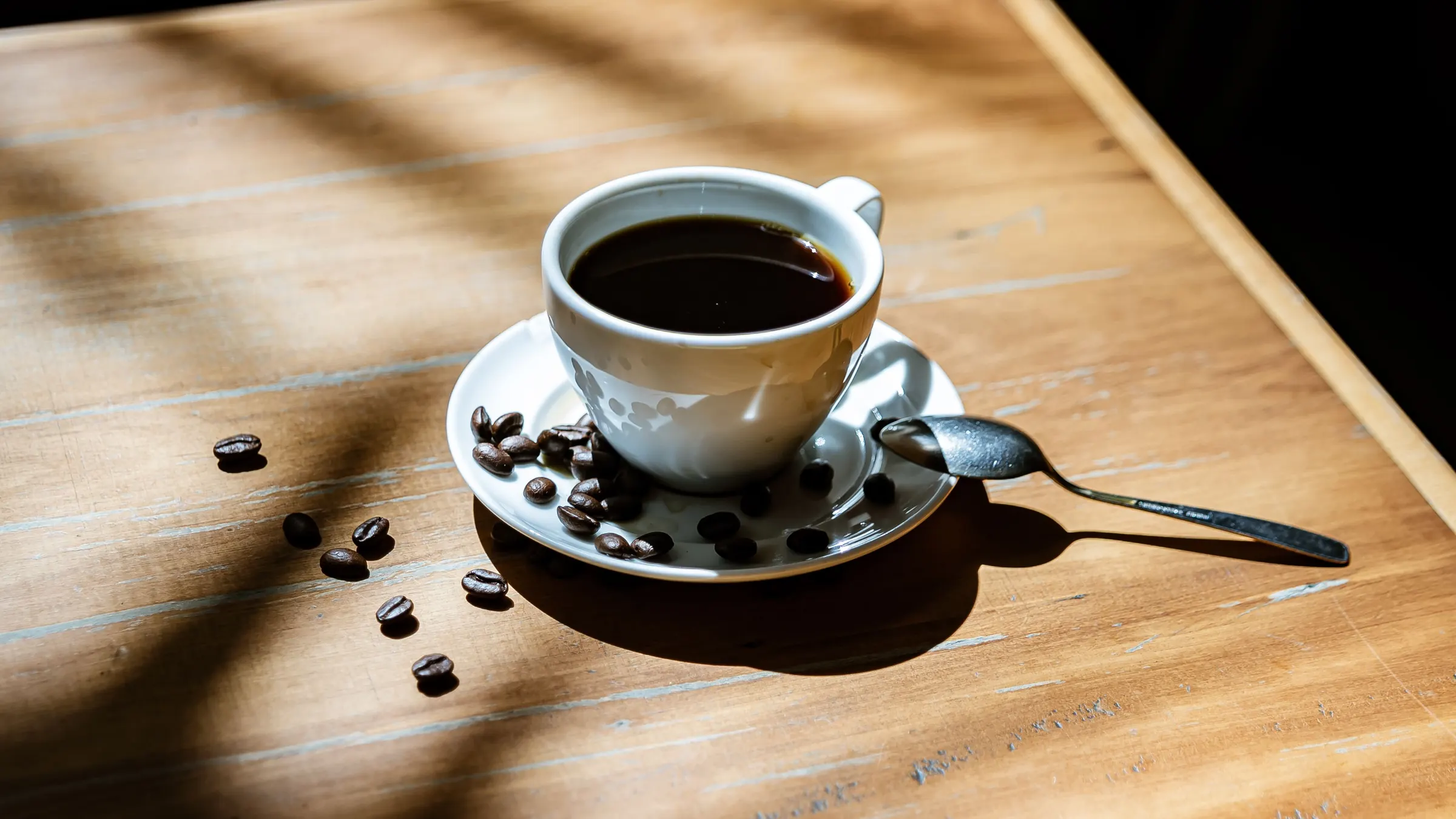 Xícara de café sobre mesa de madeira iluminada pela luz da janela, com grãos de café ao lado.